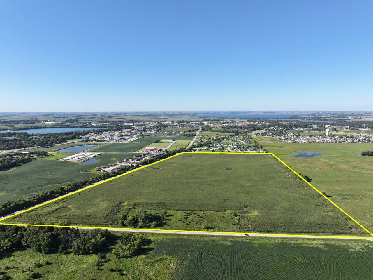 Aerial view of a vast green farmland outlined in yellow, adjacent to a small town. The landscape includes fields, buildings, and a distant lake under a clear blue sky.
