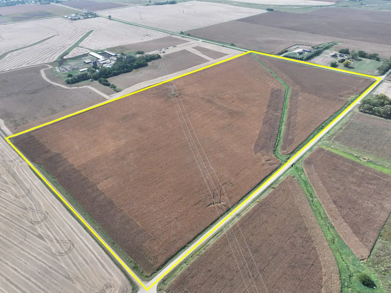 Aerial view of a large field outlined in yellow, surrounded by farmland. The field's brown soil is crossed by power lines, with a few buildings nearby.