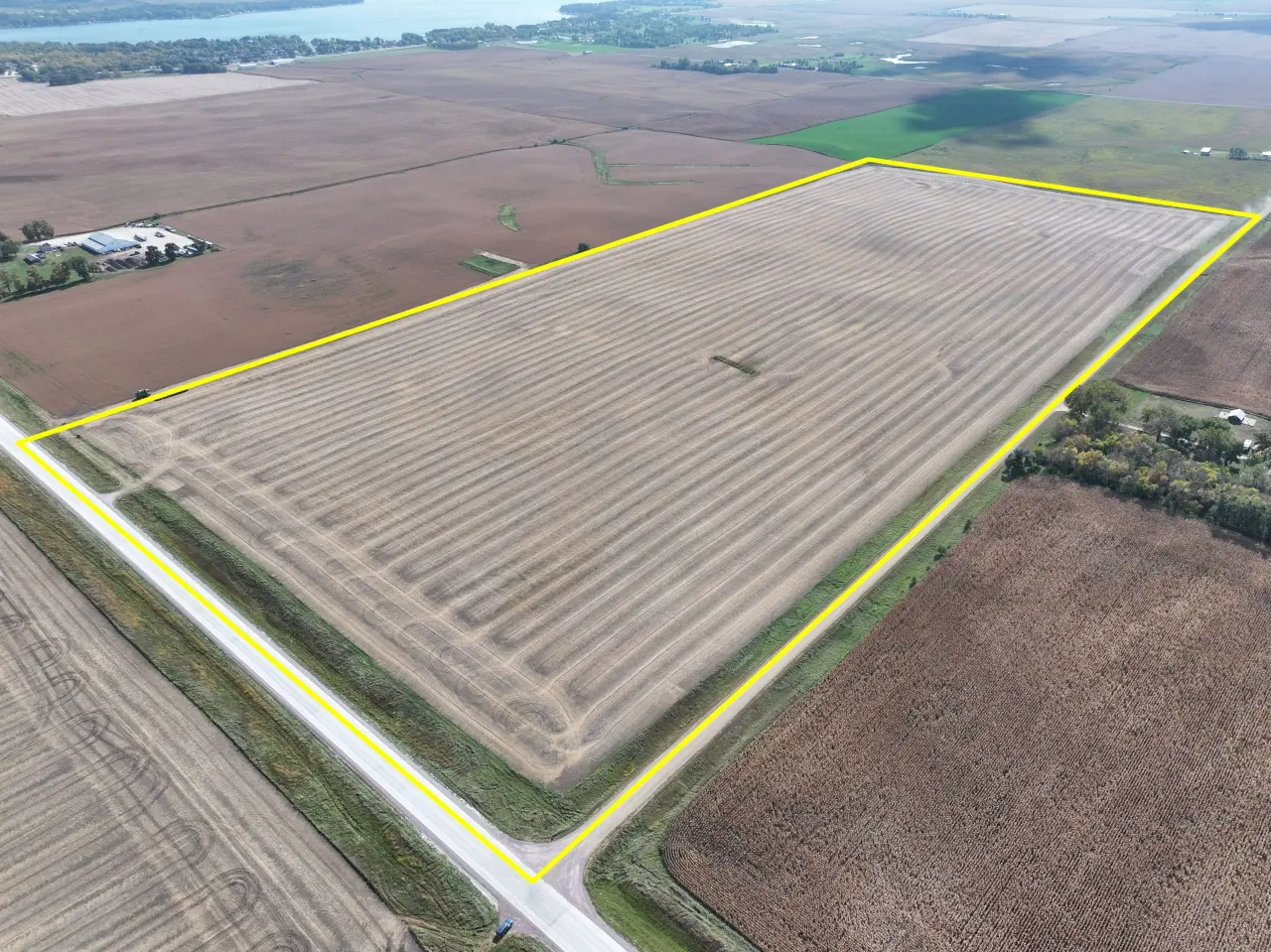 Aerial view of a large, rectangular farmland outlined in yellow. The field shows neat rows of harvested crops, surrounded by other fields and a road.