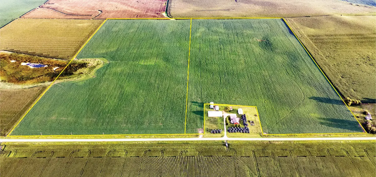 Aerial view of vast green farmlands divided by yellow lines. A cluster of farm buildings is visible in the lower right, surrounded by fields. A road runs horizontally across the bottom.