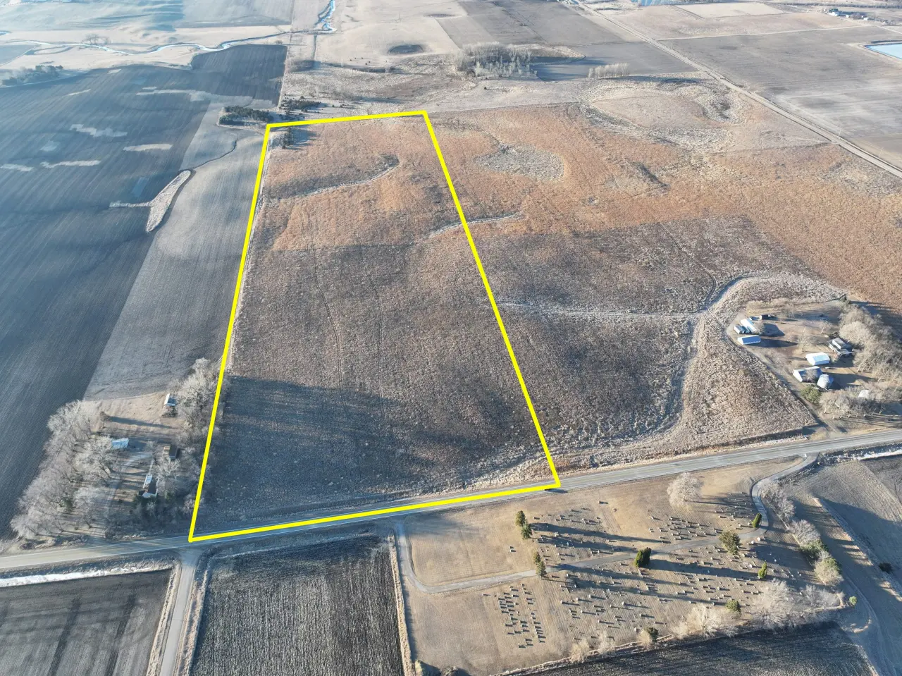 Aerial view of a large, brown field highlighted by a yellow rectangular border. Surrounding areas include barren land and sparse trees, suggesting a rural landscape.