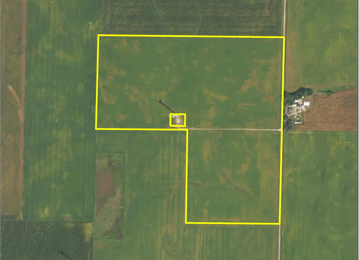 Aerial view of a vast green agricultural field outlined in yellow, showcasing rectangular plots. A small cluster of buildings is visible on the right.