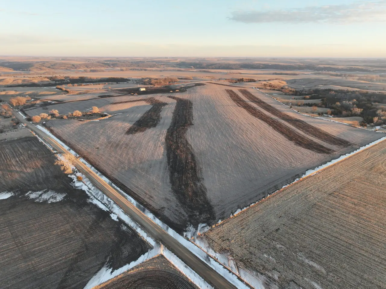 Aerial view of expansive farmland at sunrise. Fields with patches of snow and distinct plowing patterns are bordered by winding roads and bare trees. Tranquil, vast landscape.
