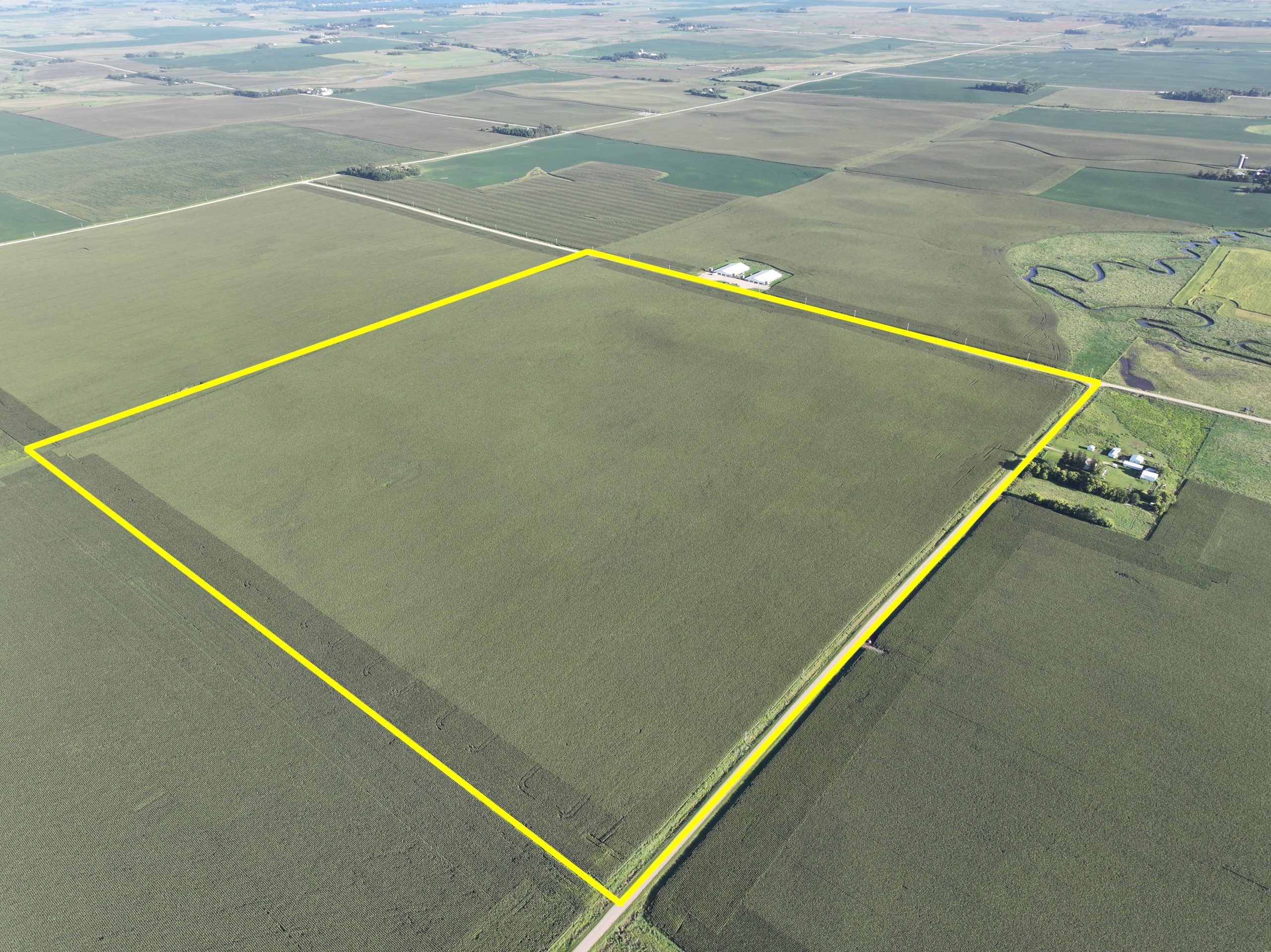 Aerial view of a large, rectangular farmland outlined in yellow, surrounded by more fields. There's a small farm building and winding stream nearby. Cloudy sky above.